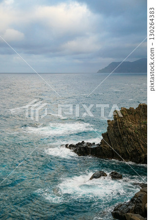 A view of sea waves crashing against a rocky cliff, with another island  130264383