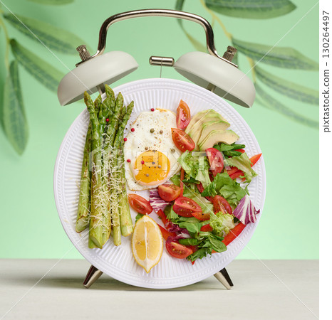Plate with asparagus, fried eggs and vegetable salad, stylized as an alarm clock on a green background. 130264497