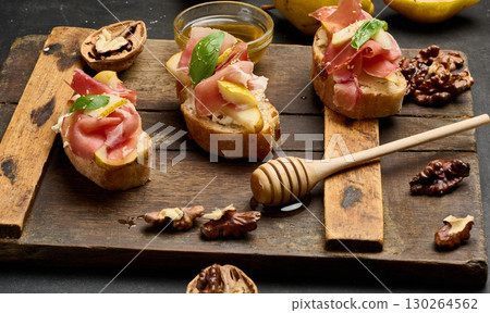 Italian bruschetta with prosciutto, pear slices, cream cheese and fresh basil, garnished with sesame seeds, served on a rustic wooden board 130264562