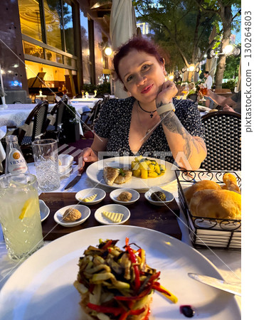 Smiling woman dining at restaurant terrace with bread, fish, potatoes, and lemonade on the table. Leisure, hospitality, and lifestyle moments in Belgrade evening atmosphere. 130264803