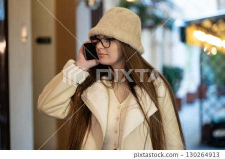 Spending a chilly afternoon in a cozy urban setting while chatting on the phone wearing a fashionable winter outfit with a fluffy hat 130264913