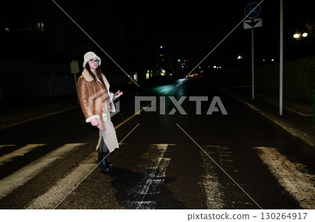 Woman crossing a quiet street at night wearing a stylish coat and boots, illuminated by distant streetlights creating a serene urban atmosphere 130264917