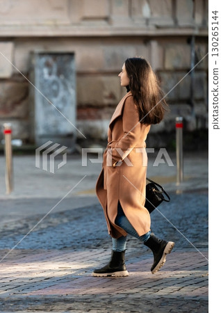 Charming stroll through the urban landscape on a sunny autumn day, highlighting fashion choices with elegant long coat and stylish boots in a lively city environment 130265144