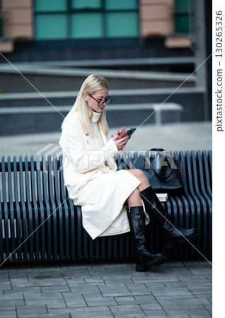 Charming woman in modern outfit enjoying smartphone while seated on urban bench amidst contemporary architecture in a bustling cityscape during the day 130265326