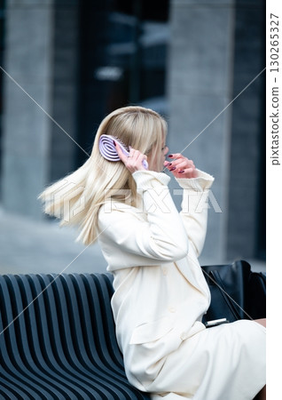 Sitting gracefully on a modern bench, a young woman with long blonde hair elegantly combs her hair on a crisp morning in a bustling urban area, embracing the moment of calm 130265327
