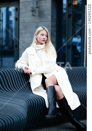 Elegant young woman in stylish winter attire relaxes gracefully on a modern bench amidst an urban backdrop during a chilly afternoon in the city Elegant young woman in stylish winter attire relaxes gracefully on a modern bench amidst an urban backdrop during a chilly afternoon in the city 130265328