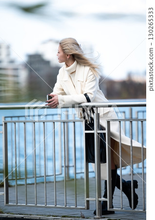 Woman in a white coat gazes thoughtfully at the water while standing by a railing on a breezy day in a modern urban setting, blending elegance with nature's beauty Woman in a white coat gazes thoughtfully at the water while standing by a railing on a breezy day in a modern urban setting, blending elegance with nature's beauty 130265333