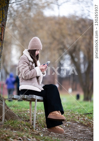 Young woman sitting on a bench in a park, absorbed in her smartphone while enjoying the calmness of a late autumn afternoon with scattered trees 130265937