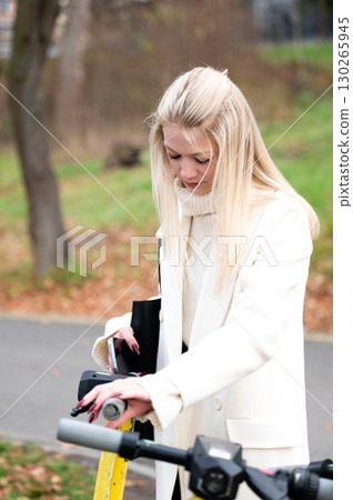 Young woman in stylish winter attire interacts with e-scooter in a scenic urban park during a crisp autumn afternoon 130265945