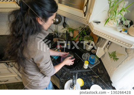 Morning melodies of water and soap a woman finds solace in washing dishes amidst a sunlit kitchen filled with potted plants and culinary tools for creating culinary delights 130266428