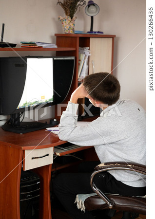 Focused youth engaged in online study while seated at a wooden desk cluttered with books and stationery in a cozy room illuminated by soft light 130266436