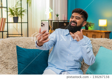 Indian man smiles taking selfie smartphone camera posts photo to social media joyful moment indoors Indian man smiles taking selfie smartphone camera posts photo to social media joyful moment indoors 130267138