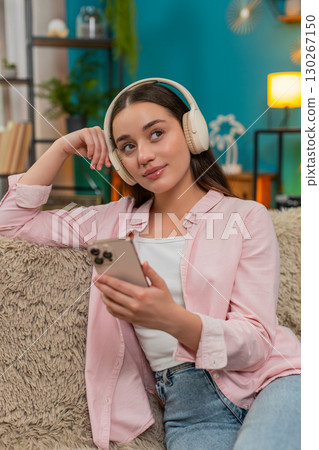 Young woman listening to podcast on sofa wearing headphones nodding while writing insightful notes 130267150