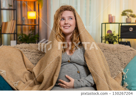Pregnant young woman wrapped in warm blanket at home gently hugging belly cozy and comforting moment 130267204