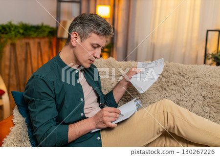 Man manages household budget writing numbers in notebook checking bills with serious focused look 130267226