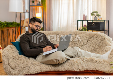Indian man writing notes from laptop into notebook at home on sofa with focused look studying online Indian man writing notes from laptop into notebook at home on sofa with focused look studying online 130267227