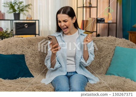 Young woman sitting on sofa anxiously checking credit card balance on smartphone worried about error Young woman sitting on sofa anxiously checking credit card balance on smartphone worried about error 130267371