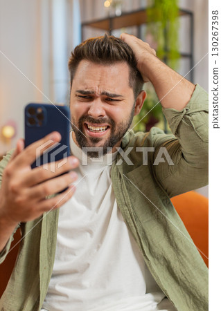 Upset displeased Caucasian man using smartphone reading bad news sitting on sofa in living room Upset displeased Caucasian man using smartphone reading bad news sitting on sofa in living room 130267398