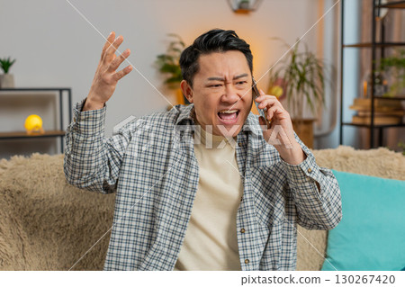Angry young Chinese man arguing on smartphone phone while sitting on couch in living room at home 130267420