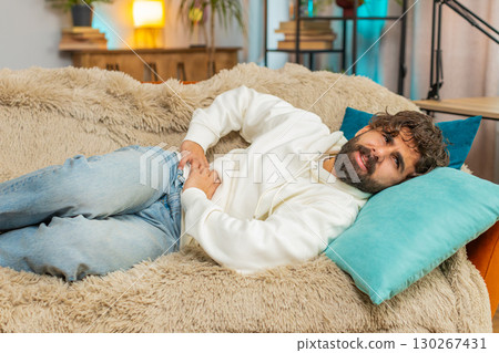 Young Indian man lying on sofa feeling sudden strong abdominal stomach ache, gastritis problem Young Indian man lying on sofa feeling sudden strong abdominal stomach ache, gastritis problem 130267431