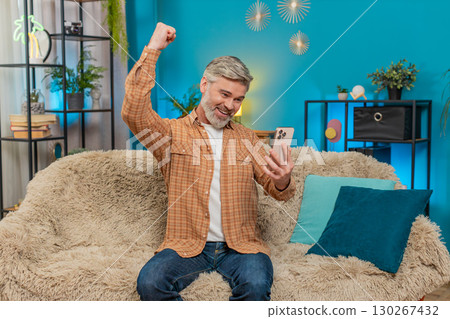 Mature man celebrating work success and online game victory while using smartphone on sofa at home 130267432