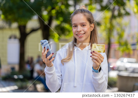Happy young woman using credit bank card and smartphone purchases online shopping on city street Happy young woman using credit bank card and smartphone purchases online shopping on city street 130267437