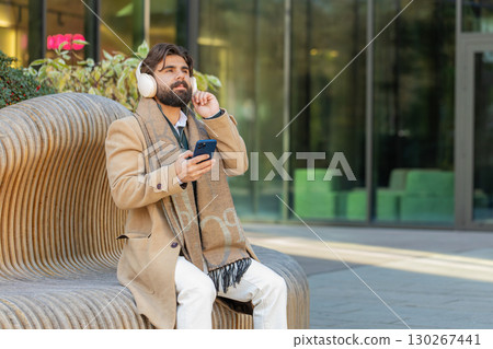 Happy Indian man in wireless headphones listening music in smartphone dancing sitting on bench Happy Indian man in wireless headphones listening music in smartphone dancing sitting on bench 130267441