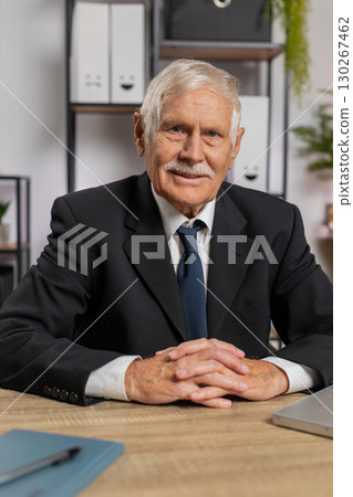 Portrait of senior businessman in suit sitting at table with laptop smiling at camera at home office Portrait of senior businessman in suit sitting at table with laptop smiling at camera at home office 130267462