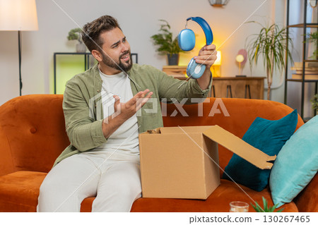Unhappy man unpacking carton box feeling dissatisfied with getting damaged wireless headphones home Unhappy man unpacking carton box feeling dissatisfied with getting damaged wireless headphones home 130267465