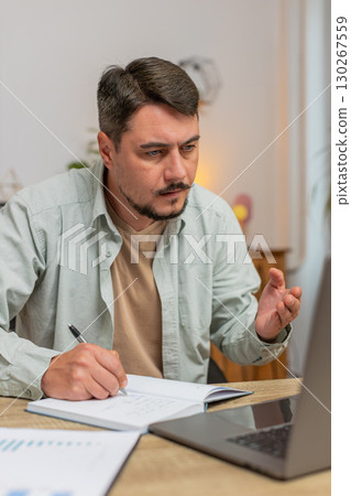 Young man writing down notes while attending online office meeting on laptop at home office desk 130267559