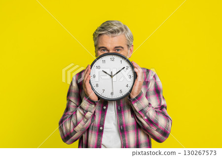 Mature man hiding behind large office clock, playful peek, cautious glance, time awareness humor 130267573