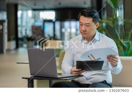 Chinese male entrepreneur in shirt with laptop analyzing graph documents reports in office lobby 130267603
