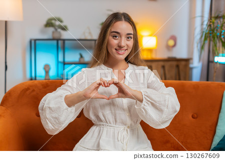 Portrait of happy Caucasian young woman makes symbol of love showing heart sitting on sofa at home Portrait of happy Caucasian young woman makes symbol of love showing heart sitting on sofa at home 130267609