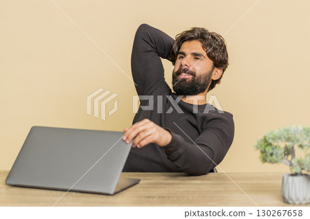 Happy young Indian man closing laptop PC after finishing work at table against beige background 130267658