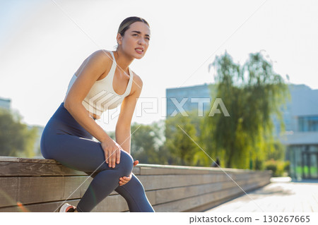 Athletic jogger woman in sportswear with knee pain after workout in urban city park on sunny day Athletic jogger woman in sportswear with knee pain after workout in urban city park on sunny day 130267665