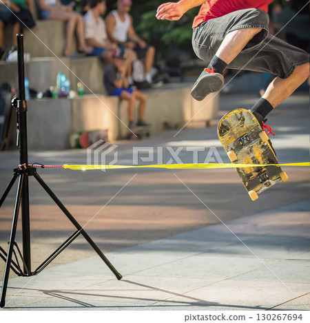 Skateboarder Mid-Jump Over Rope. Person performing jump on skateboard over stretched yellow tape, street sport, street competition. Concept of skateboarding, urban tricks, extreme sport 130267694