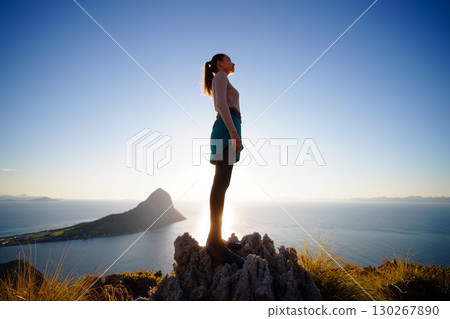 Woman Standing on a Rock Overlooking the Ocean at Sunset Near a Mountainous Island 130267890