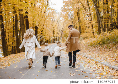 Young man, woman and their children walking outside wearing beige coats. Blond mother, brunette father, little girl and boy holding hands. Parents wearing coats, children - hats. 130267931