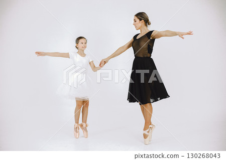 Little girl ballerina with her mother on white background. Brunette girl wearing white ballet suit and adult woman black dress. Two female dancer dancing together in a pointe shoes. 130268043