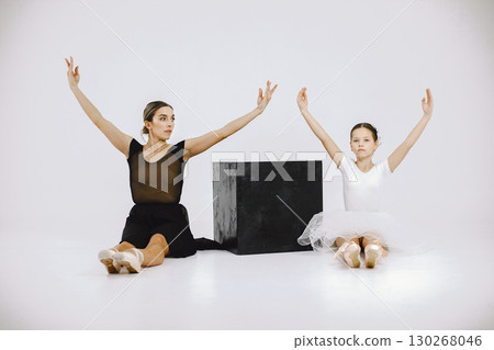 Little ballerina with her mother on white background. Brunette girl wearing white ballet suit and adult woman black dress. Two female dancer sitting near black cube. Little ballerina with her mother on white background. Brunette girl wearing white ballet suit and adult woman black dress. Two female dancer sitting near black cube. 130268046