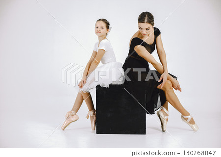 Little ballerina with her mother on white background. Brunette girl wearing white ballet suit and adult woman black dress. Two female dancer sitting on a black cube. Little ballerina with her mother on white background. Brunette girl wearing white ballet suit and adult woman black dress. Two female dancer sitting on a black cube. 130268047