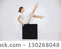 Little ballerina on white background. Brunette girl wearing white ballet suit. Girl sitting on black cube. 130268048