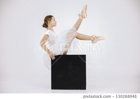 Little ballerina on white background. Brunette girl wearing white ballet suit. Girl sitting on black cube. 130268048