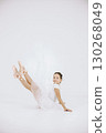 Little ballerina on white background. Brunette girl wearing white ballet suit. Girl sitting on a floor. 130268049