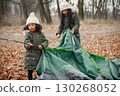Two little black girls in tent camping in the forest. Two little sisters are puting up a tent in autumn forest. Black girls wearing khaki coats and beige hats. 130268052