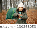 One little black girl in tent camping in the forest. Girl is holding a thermos in her hands. Black girl wearing khaki coat and beige hat. 130268053