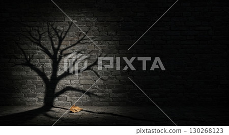 A dramatic shadow of a leafless tree is cast upon a dark brick wall, with a single withered leaf resting on the cracked ground below, creating a stark and moody scene. 130268123