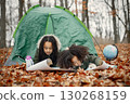 Beautiful little black girls in tent camping in the forest. Two little sisters lying in a tent in autumn forest and looking on a map. Black girls wearing khaki coats. 130268159