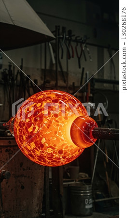 A glowing orange and red glass orb with a textured surface is being shaped on a blowpipe in a dark glassblowing workshop, illuminated by its own intense heat. A glowing orange and red glass orb with a textured surface is being shaped on a blowpipe in a dark glassblowing workshop, illuminated by its own intense heat. 130268176