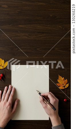 A person's hands are positioned on a dark wooden desk, with one hand holding a fountain pen above a blank cream paper, flanked by colorful autumn leaves and red berries. 130268219